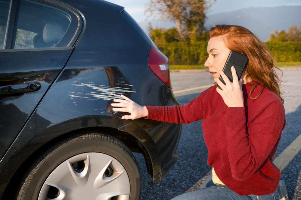 Red haired woman on phone looking at large scratch on side of car
