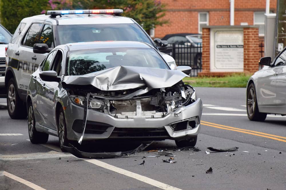 A car with significant front end damage from an accident, with a police car behind it