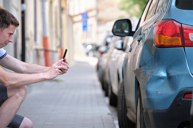 Driver in shorts and tshirt crouching to take a picture of car damage