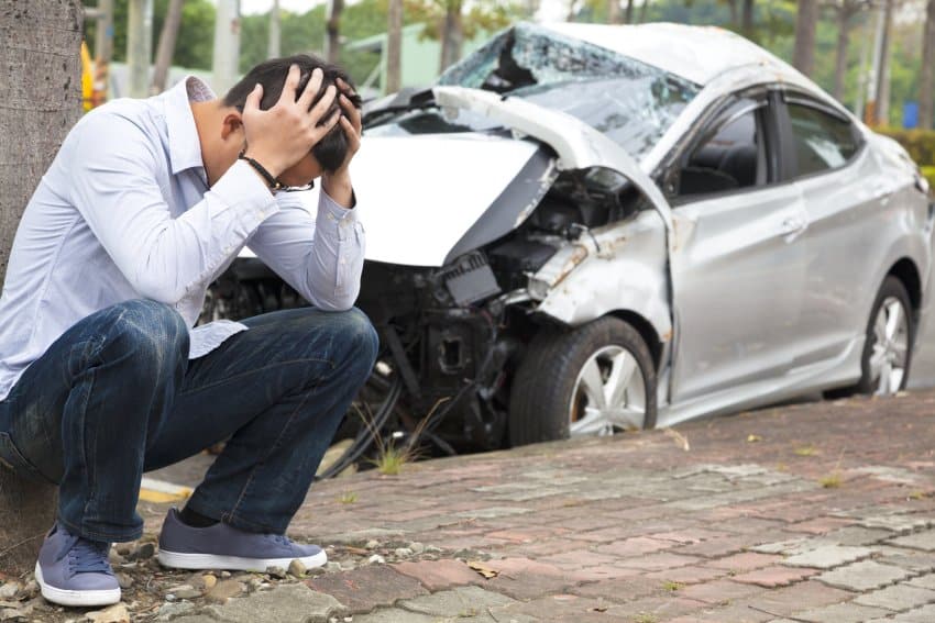 Male driver squatting on ground holding head in hand after crashing gray car front collision accident