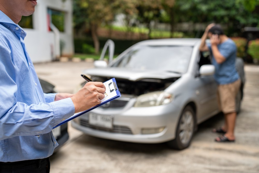 Insurance handler taking notes on clipboard frustrated driver in blue shirt standing beside gray car in background