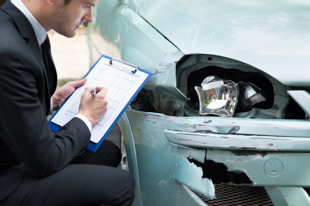 Insurance agent crouched with clipboard beside damaged turquoise car filing report