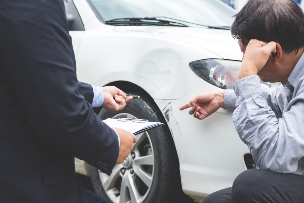 Insurance agent and car accident victim inspecting white car damage to wheel and scratched bumper
