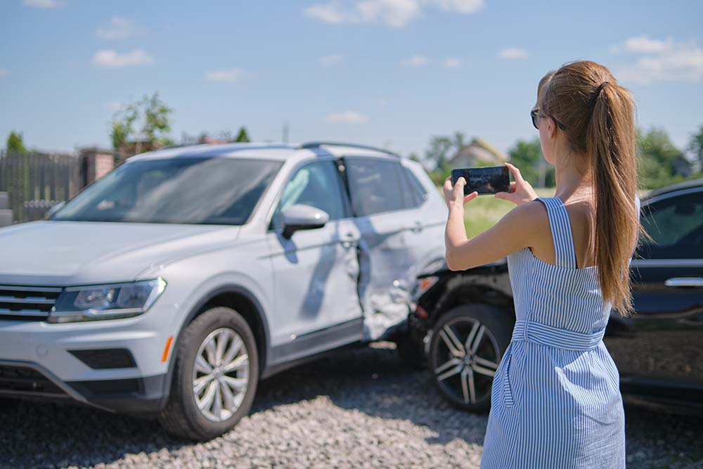 Young woman in blue dress taking picture of side damage to car after crash