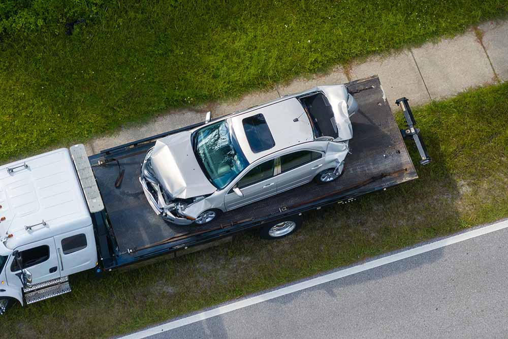 A white car with front and rear end crumpled, on the bed of a tow truck on the side of the road after an accident.