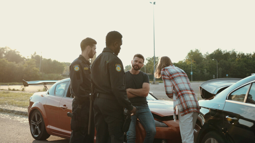 Drivers, witnesses, and a police officer speak with one another trying to resolve a car crash situation. In the background, a red sports car’s hood is crumpled from rear-ending a black car in front of it.