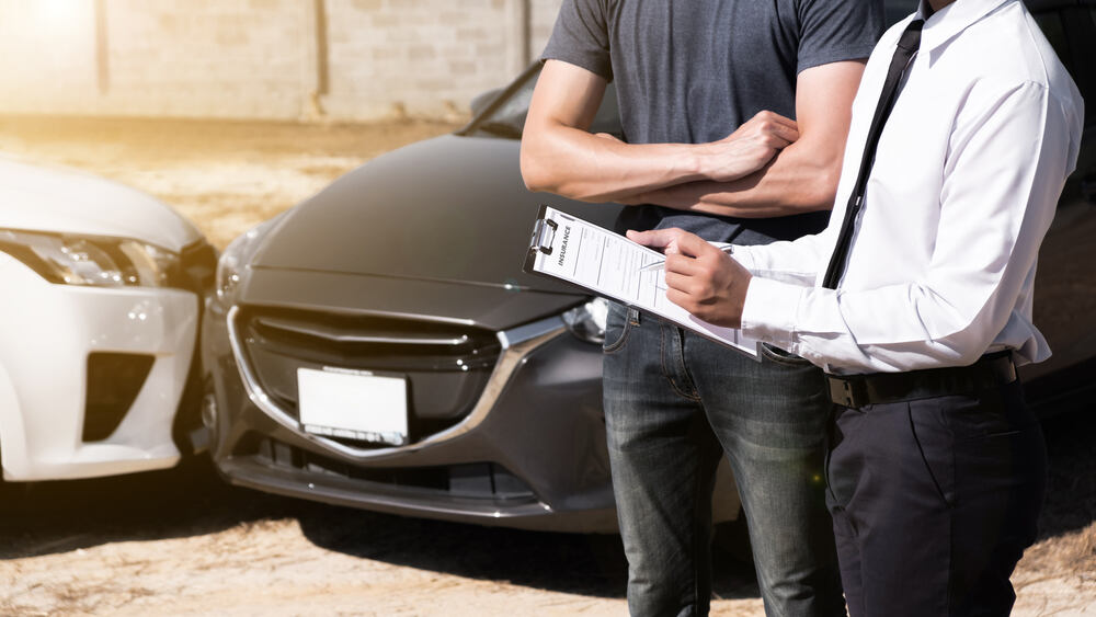 An insurance agent stands in front of two cars that have front-end collided. He appears to be filling out a report while a person stands beside them, arms crossed. 