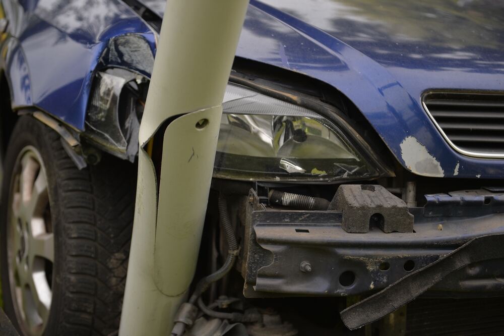 A blue car, it’s light and bumper damaged after running into a lamp post which is slightly leaning, and damaged, into the broken front end of the car. 