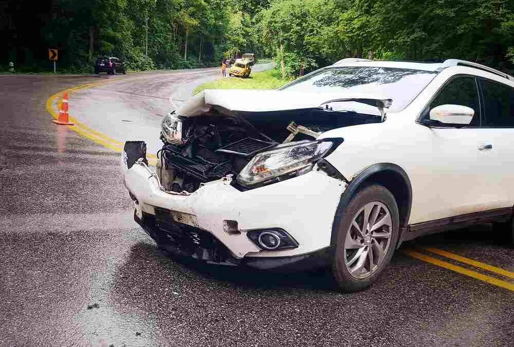 A white SUV resting the wrong way across a no-pass double yellow line, it’s front end is rumpled from a collision and the front bumper appears to be barely hanging on.