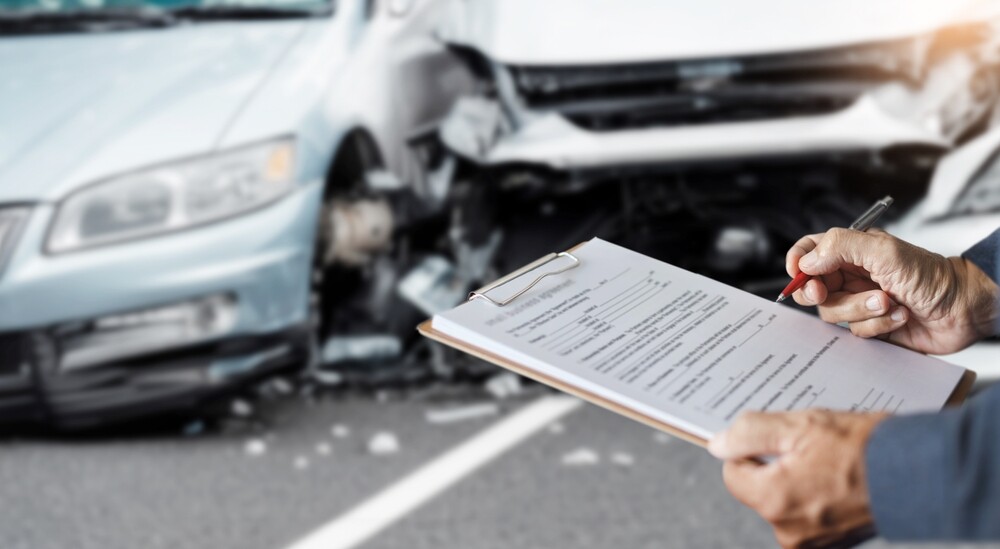 An insurance agent in focus at the scene of a collision between two light-colored cars in the background, softly blurred. The agent is filling out a report.