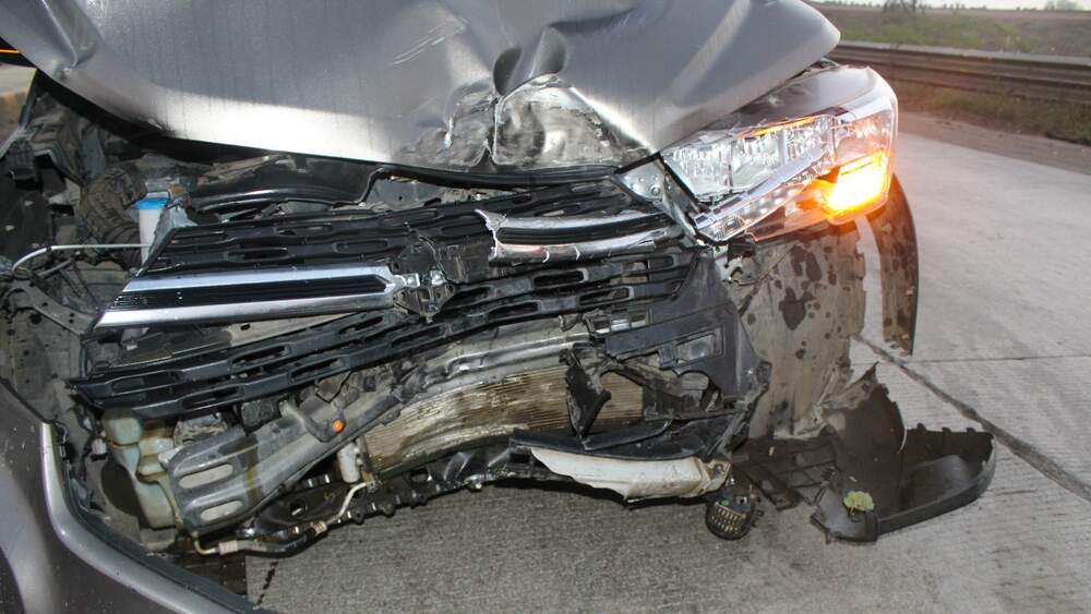 The front end of a dark colored vehicle, crumpled inward, with one turn signal light still working.