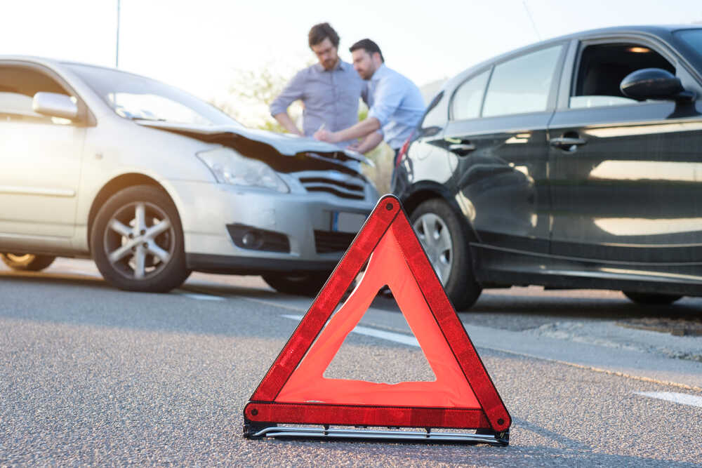 Two men in the aftermath of a collision, a white car and dark black car show light damages, while the two men fill out an accident report on the hood of the white car.